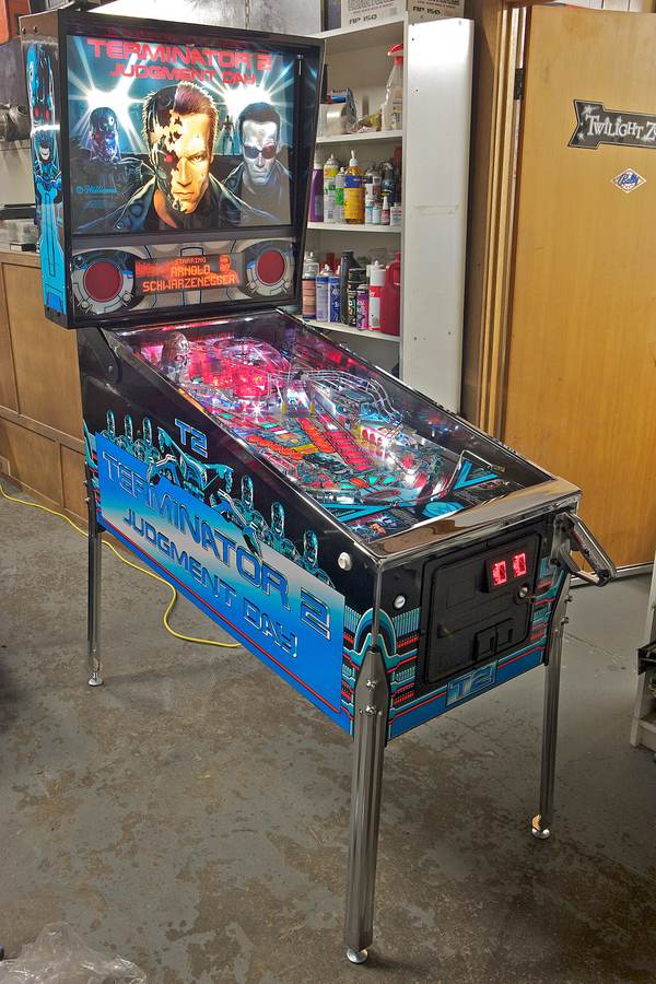 A restored Terminator 2: Judgment Day pinball machine with a lit-up backglass and blue cabinet art, standing in a workshop